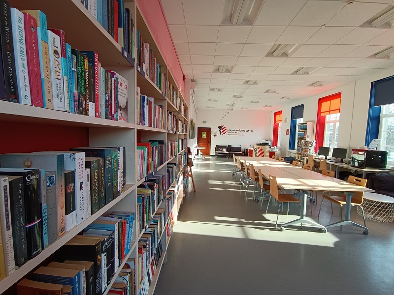 Bibliotēkas telpa ar grāmatu plauktiem, galdiem, datoriem tālu fonā un uzrakstu ASV Informācijas centrs Daugavpils