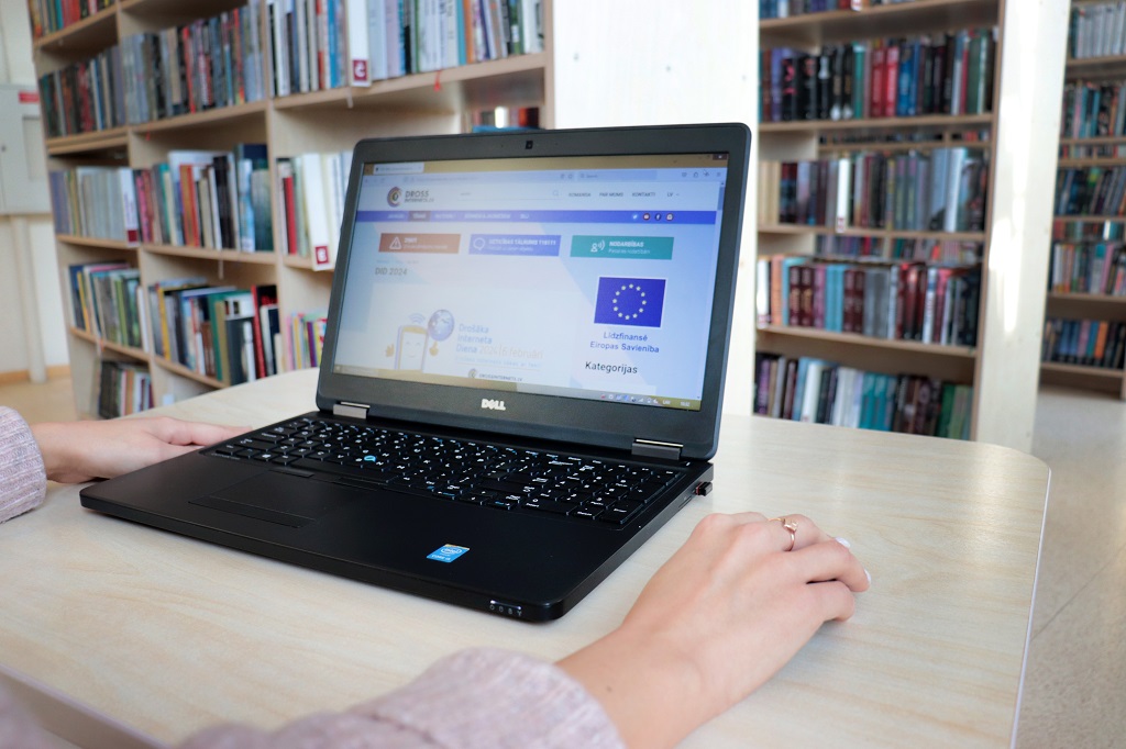 Meitene sēž pie galda uz kura novietots portatīvais dators ar atvērtu mājaslapu "Drošs internets" un fonā grāmatu plaukti