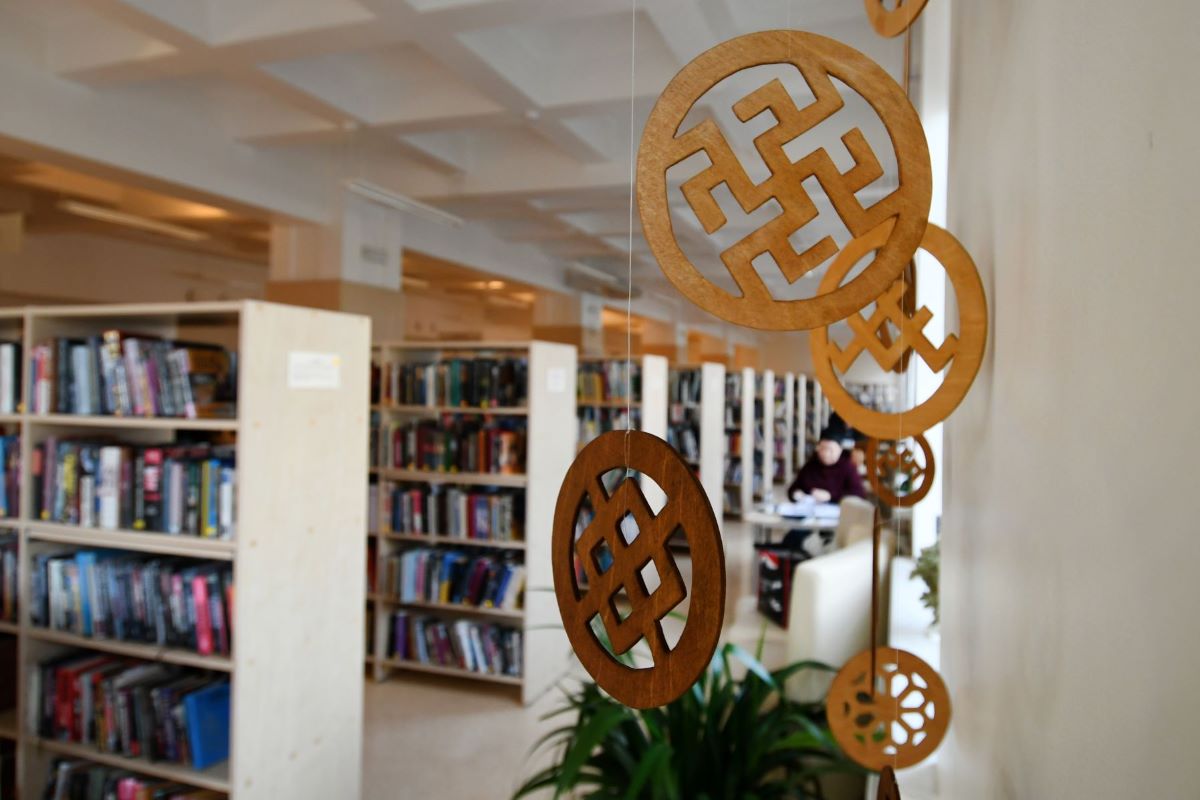 Patriotu dienas 2023 Ilustratīvs attēls ar piekārtām Latvju zīmēm un fonā bibliotēkas telpa ar grāmatu plauktiem un lasītājiem, kuri lasa grāmatas pie galdiem