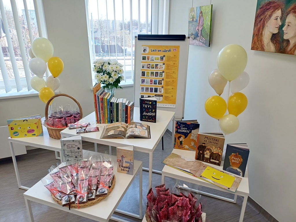 Mājīgs stūrītis ar krāsainu bērnu grāmatu izstādi uz baltiem galdiem. Grozi ar dāvanu iesaiņojumiem, dzelteni un balti baloni, kā arī informatīvs plakāts papildina dzīvespriecīgo noformējumu.