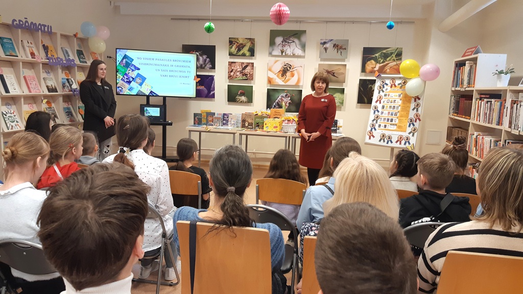 Sieviete sarkanā kleitā uzstājas sēdošai auditorijai bibliotēkā. Izstādītas grāmatas un aizmugurē fotogrāfiju izstāde ar dzīvnieku fotogrāfijām, fonā redzams prezentācijas ekrāns.