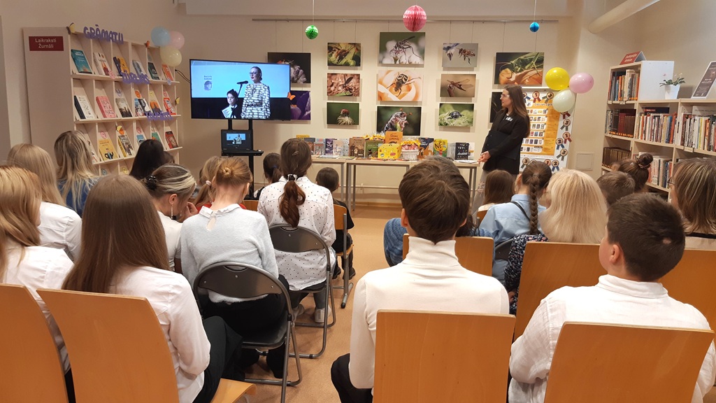 Bērnu un pieaugušo grupa sēž bibliotēkā un klausās sievietes stāstījumā. Gar sienām ir grāmatu plaukti, un telpu rotā krāsaini baloni.