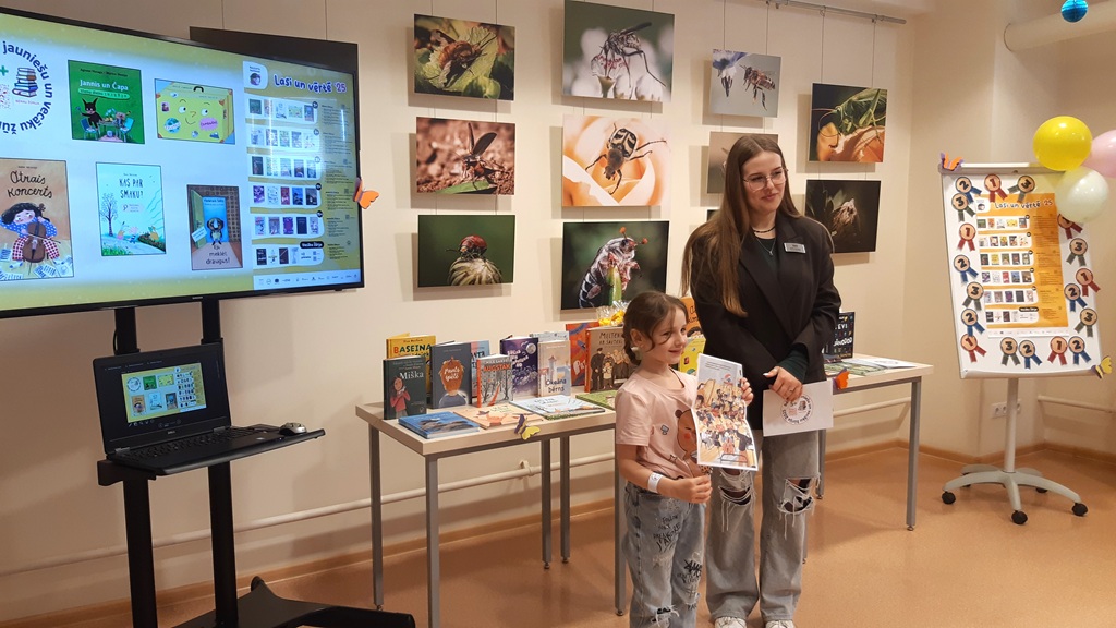 Sieviete un bērns smaidot tur grāmatu bibliotēkas telpā, pie kuras sienas ir dzīvnieku fotogrāfijas. Redzama grāmatu izstāde un prezentācijas ekrāns.