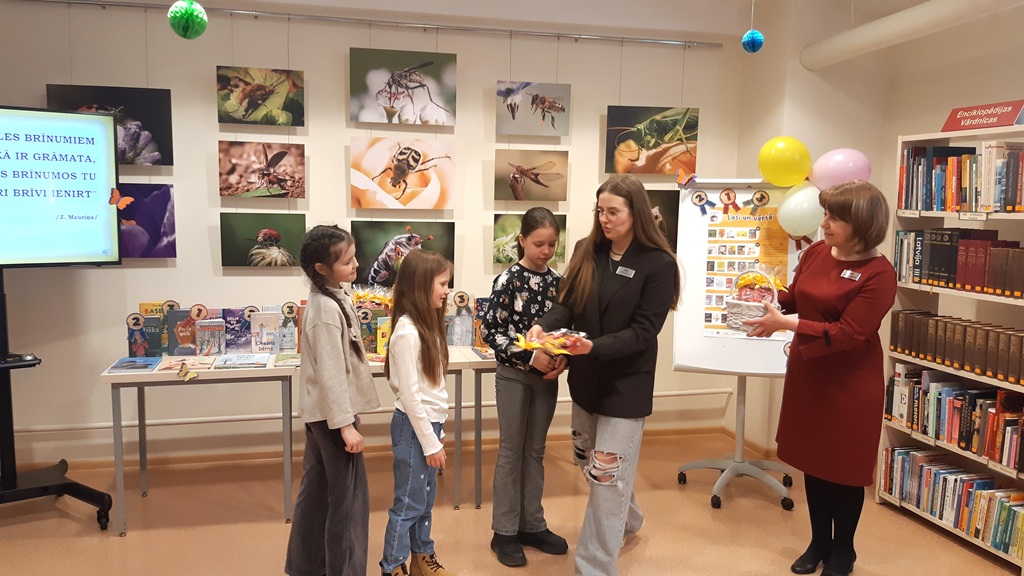 Trīs bērni stāv blakus divām sievietēm bibliotēkā. Viena no sievietēm sniedz dāvanu starp krāsainām, dzīvnieku tematikai veltītām fotogrāfijām.