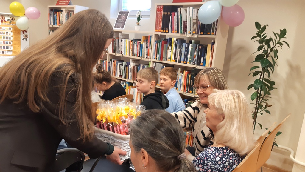 Sieviete bibliotēkā dala kārumus, apkārt sēž smaidīgi dalībnieki. Grāmatu plaukti, krāsaini baloni un silta, aicinoša atmosfēra raksturo šo ainu.