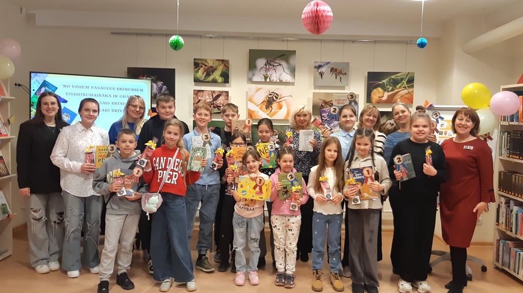 Bērnu un pieaugušo grupa pozē bibliotēkā, smaidot un turot rokās grāmatas un diplomus. Telpa ir dekorēta ar krāsainiem baloniem, radot priecīgu gaisotni.