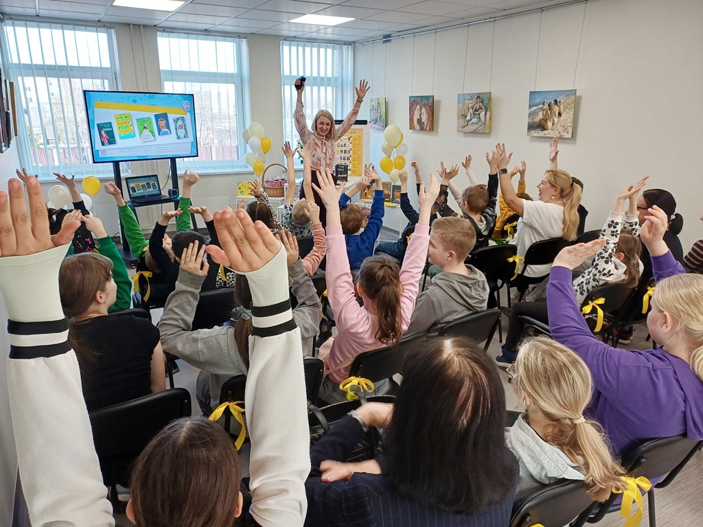 Dzīvīga bibliotēkas telpa ar sēdošiem bērniem, kuri sajūsmā ceļ rokas pret smaidīgu sievieti telpas priekšā. Baloni un ekrāns papildina svētku noskaņu.