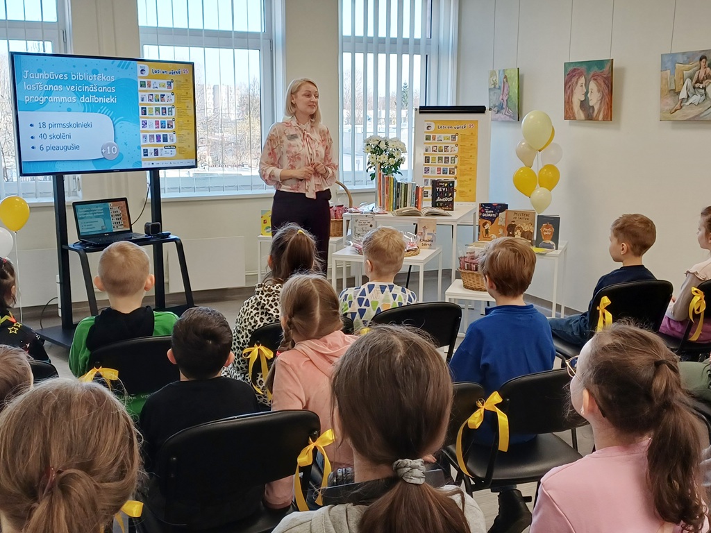 Sieviete stāv gaišas telpas priekšā un sniedz prezentāciju sēdošiem bērniem, kuriem pie krēsliem piespraustas dzeltenas lentes. Slaidā redzami dati par dalībnieku skaitu.
