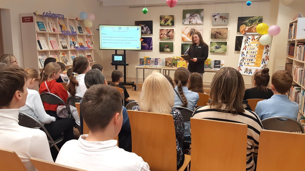 Sieviete uzstājas bērnu un pieaugušo auditorijai bibliotēkā. Blakus grāmatu plauktiem redzams ekrāns ar tekstu. Telpa ir krāsaina un saistoša.