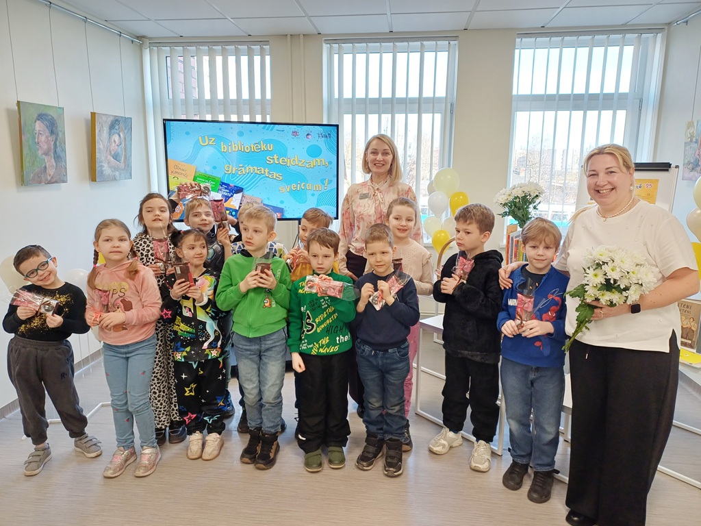 Smaidīgu bērnu grupa un divi pieaugušie ar dāvanām rokās gaišā telpā. Ekrāns un baloni fonā liecina par svinīgu pasākumu.