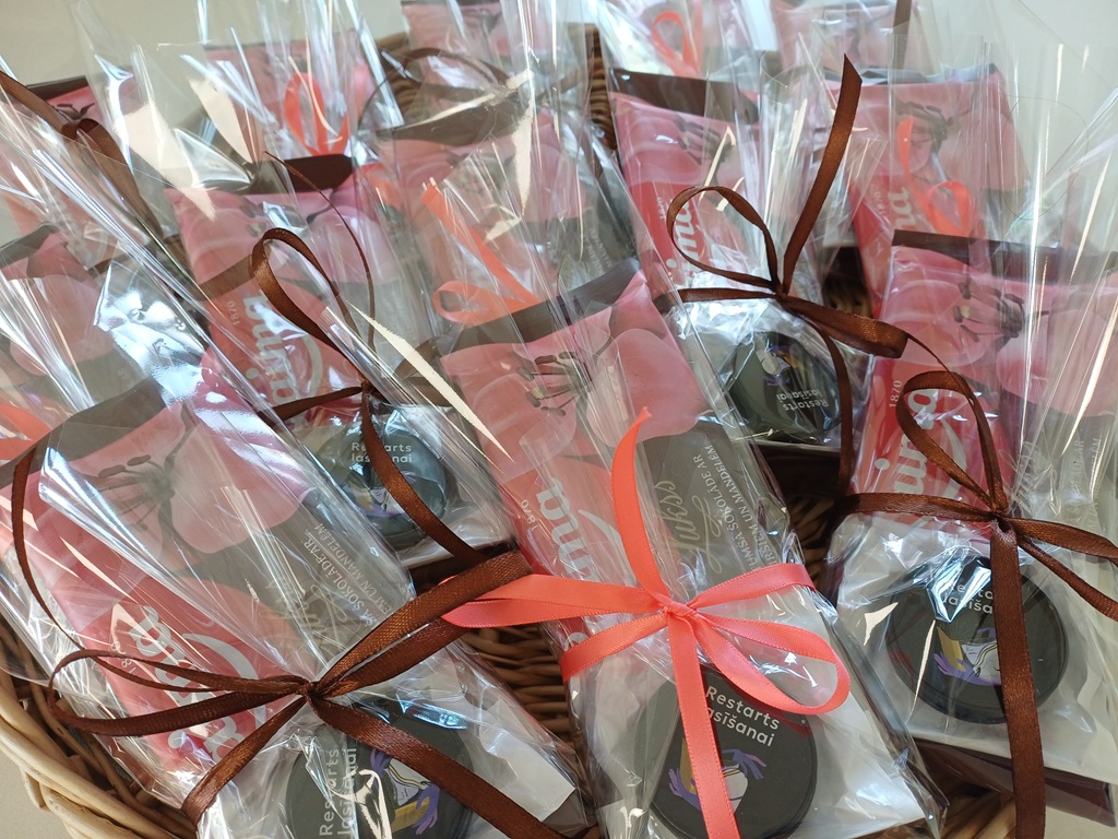 Grozs, kas pildīts ar dāvanu saiņojumiem, kuros atrodas šokolāde un viens priekšmets ar uzrakstu 'Restarts lasīšanai'. Iesaiņojumi pārsieti ar tumši sarkanīgām un rozā lentītēm, radot svinīgu un dāvanām bagātu iespaidu.