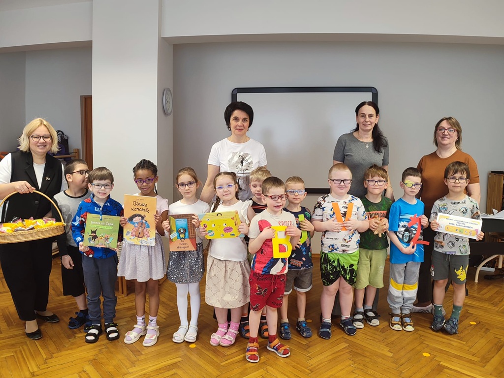 Smaidīgu bērnu grupa ar grāmatām un burtiem rokās stāv nodarbību telpā kopā ar četriem pieaugušajiem. Vienam no pieaugušajiem rokās ir grozs. Gaisotne ir priecīga un izzinoša.