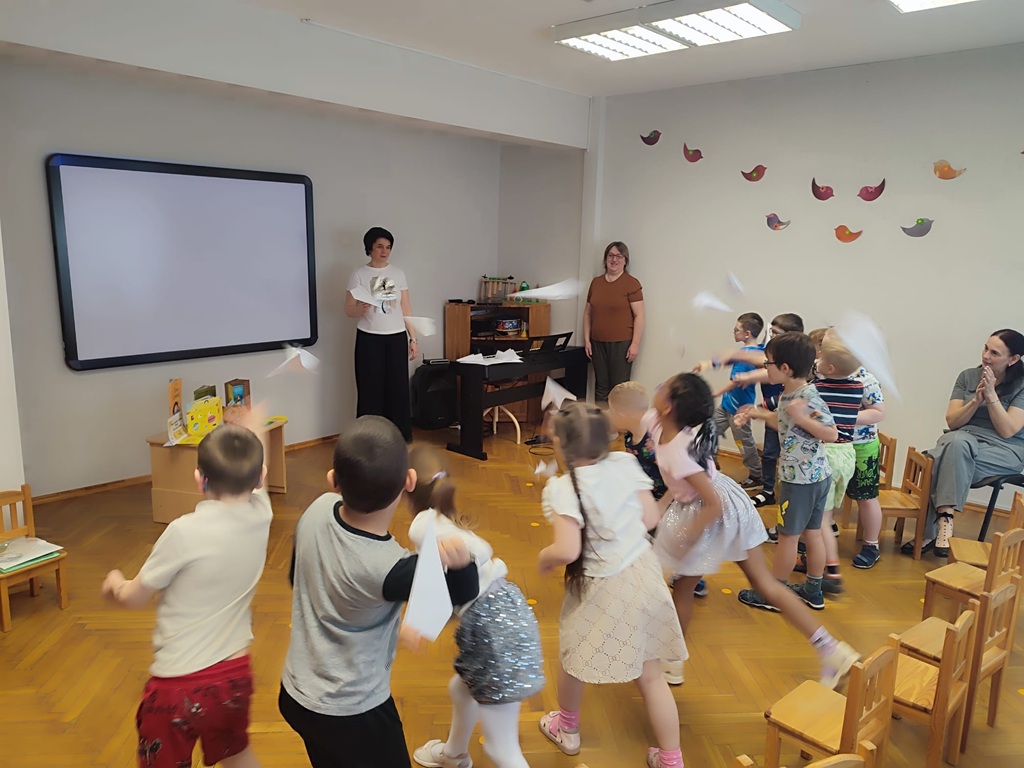 Bērni klasē priecīgi laiž papīra lidmašīnas divu pieaugušo uzraudzībā. Telpā ir koka grīda, liels ekrāns un krāsainas putnu uzlīmes pie sienas.
