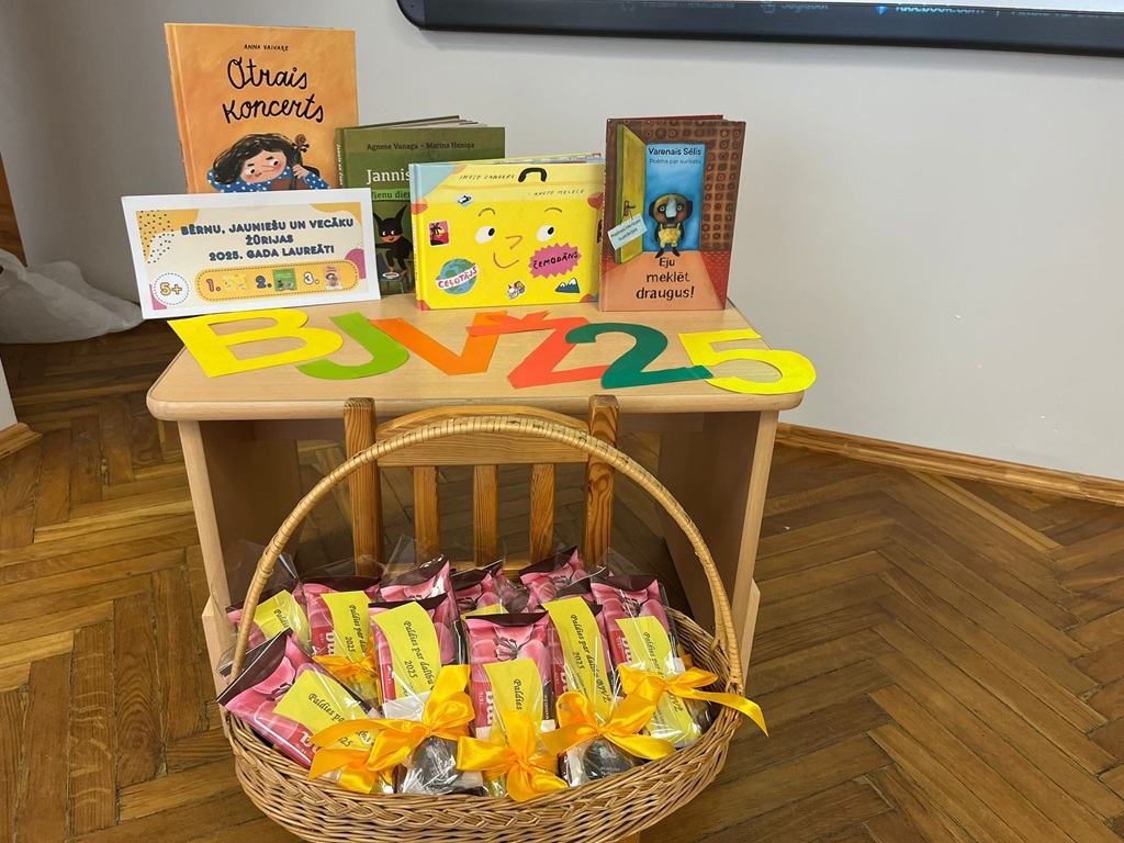 Uz koka galda izvietotas bērnu grāmatas, krāsaini burti 'BJVŽ 25' un grozs ar kārumiem, kas apsieti ar dzeltenām lentēm. Noskaņa ir svinīga un izzinoša.