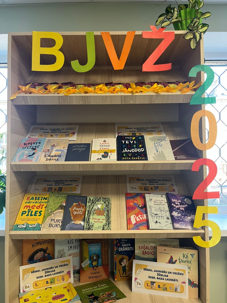Grāmatu plaukts, kura augšpusē ir burti "BJVŽ", bet sānos vertikāli izvietots gada skaitlis "2025". Plauktā atrodas dažādas bērnu grāmatas krāsainos vākos.