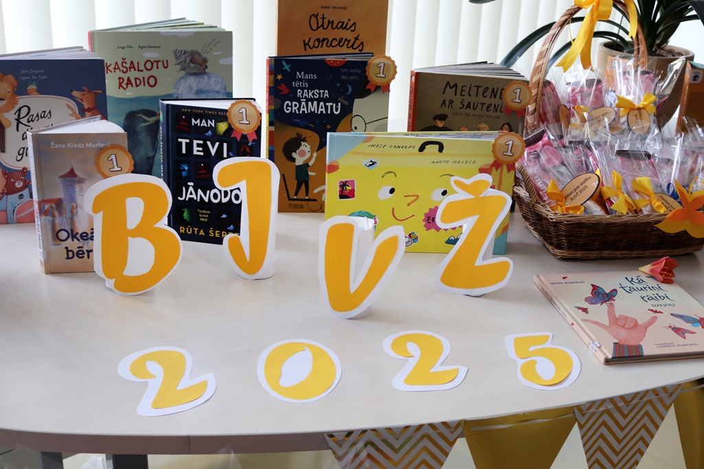 Uz galda ir izvietotas krāsainas, godalgotas grāmatas ar pirmās vietas etiķetēm. Dzelteni burti veido "BJVŽ 2025". Grozs ar dekoratīviem priekšmetiem piešķir svētku noskaņu.