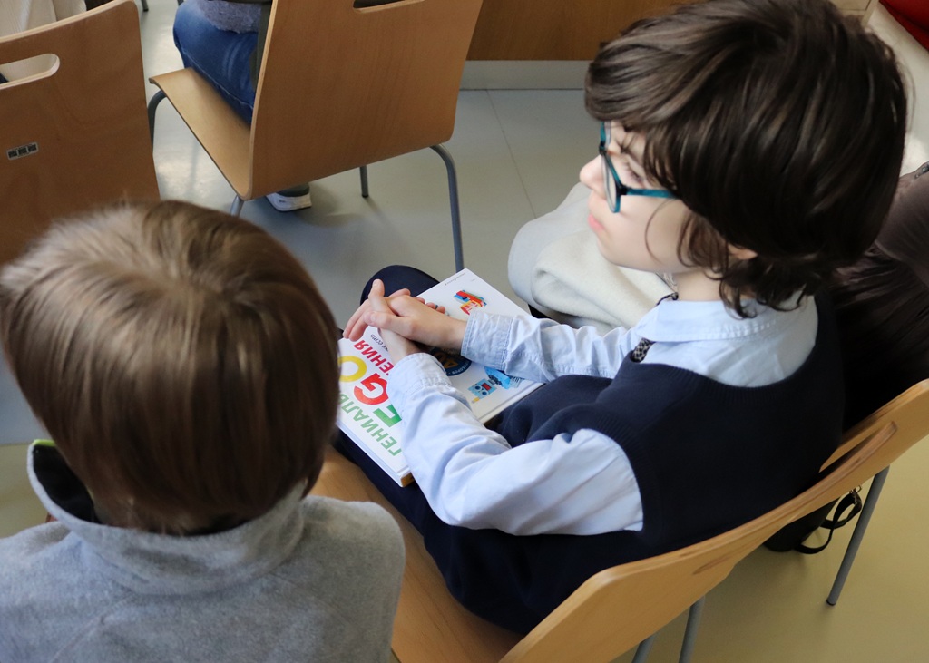 Divi bērni sēž, viens tur krāsainu grāmatu. Viņu koncentrētās sejas izteiksmes un klases gaisotne rada mierīgu, uzmanīgu atmosfēru.