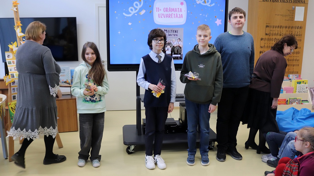 Četri skolēni smaidoši stāv pie ekrāna, uz kura drukāts "11+ Grāmata Uzvarētāja", klasē radot jautru un svinīgu atmosfēru.