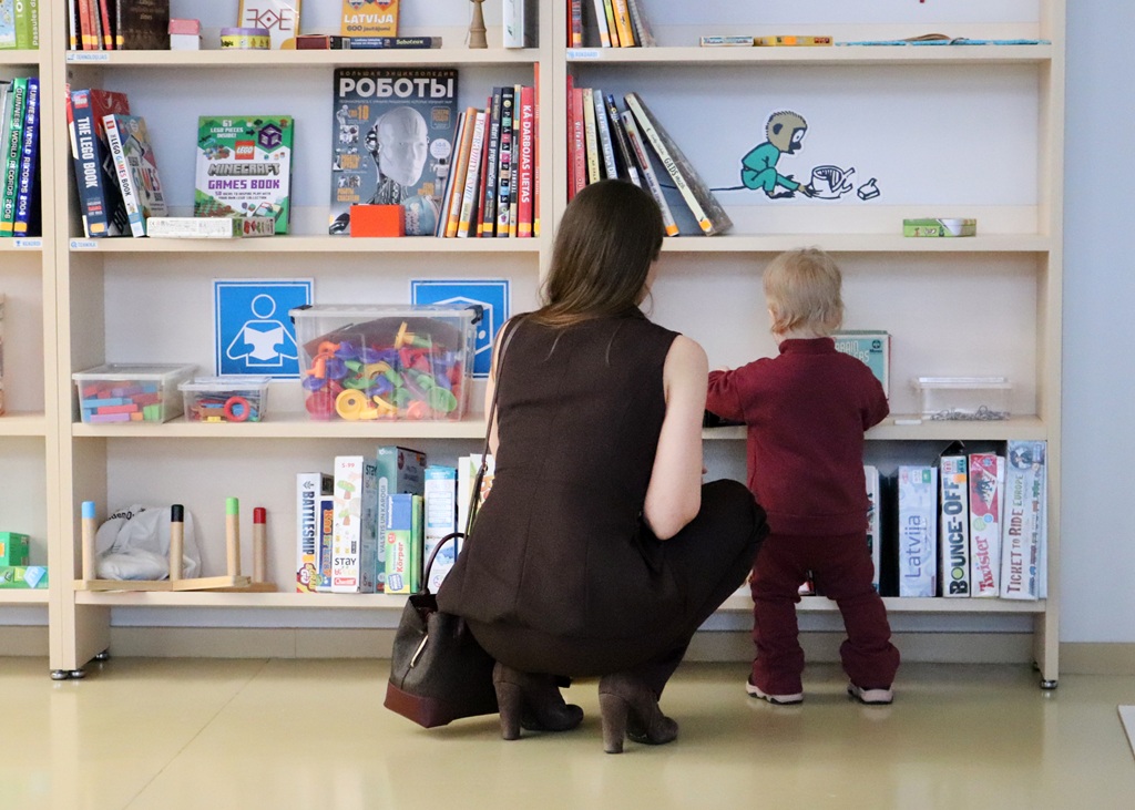 Sieviete un mazulis stāv pie grāmatu plaukta, kas pilns ar krāsainām grāmatām un rotaļlietām. Sieviete pietupusies vēro, kā bērns visu izpēta.