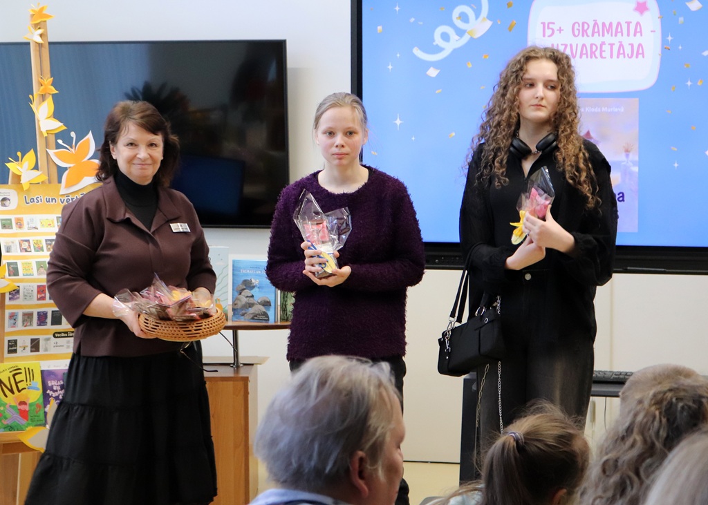 Trīs cilvēki stāv pasākuma telpās. Divi tur dāvanu maisiņus, bet viens tur grozu ar našķiem. Spilgts ekrāns fonā rada svinīgu noskaņu.