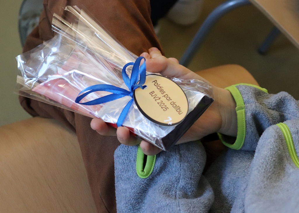 Bērna rokā ir iesaiņota dāvana, kas sasieta ar zilu lentīti un uz kuras ir piestiprināta birka ar tekstu.