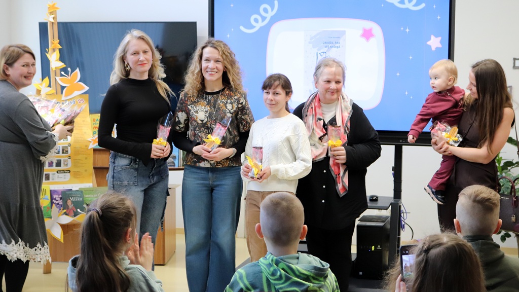 Piecu pieaugušo grupa, četri no viņiem tur dāvanu maisiņus, smaidot stāv auditorijas priekšā. Sieviete, turot rokās bērnu, ir labajā pusē. Vide ir dzīvespriecīga, un aizmugurē ir ekrāns ar krāsainiem attēliem.