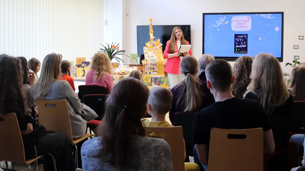 Sieviete rozā žaketē uzrunā ieinteresētu auditoriju, tostarp bērnus, gaišā telpā. Krāsains displejs un ekrāns pastiprina dzīvespriecīgo atmosfēru.