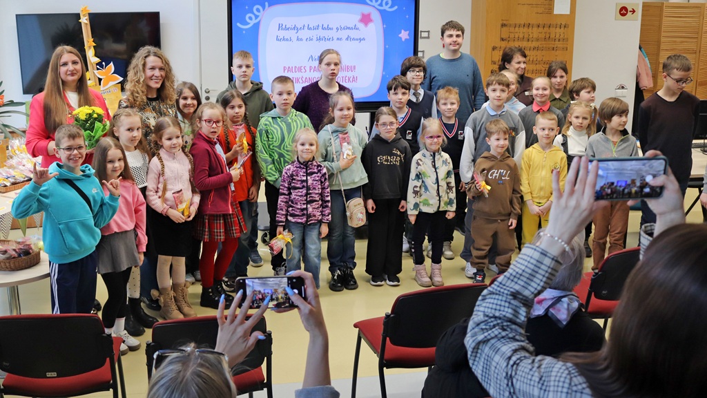 Bērnu grupa un divi pieaugušie smaida un pozē fotogrāfijai klasē. Daži cilvēki priekšplānā iemūžina mirkli ar viedtālruņiem.