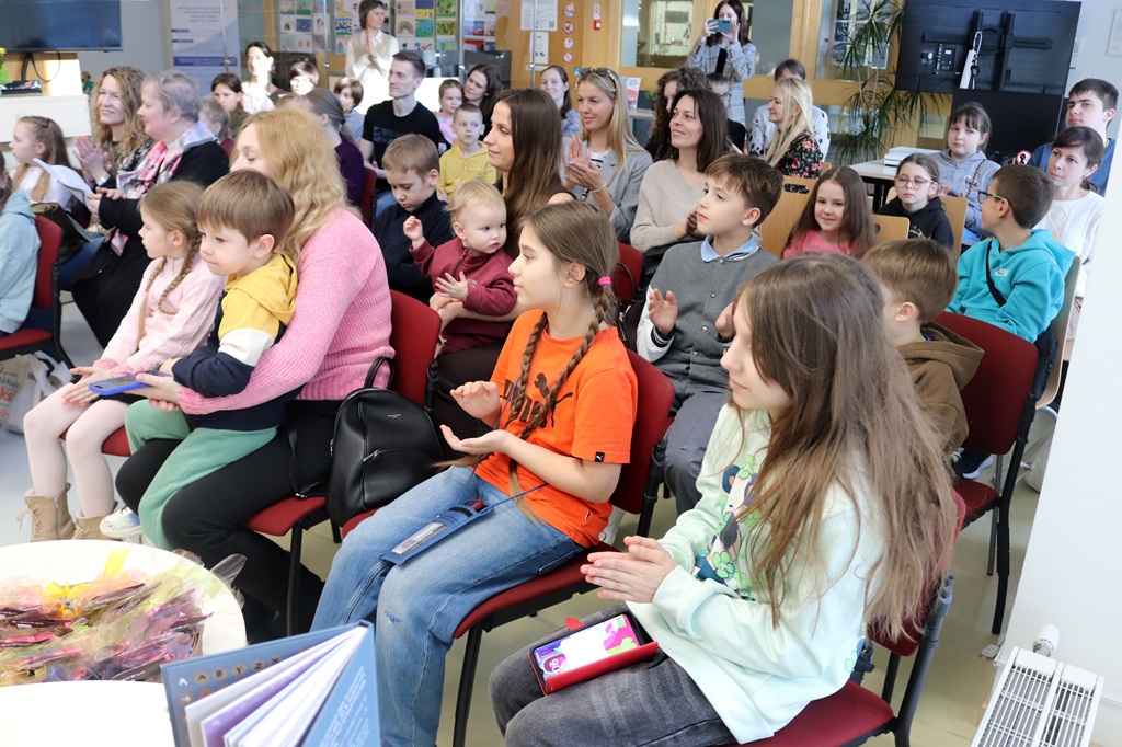Bērnu un pieaugušo auditorija sēž uz sarkaniem krēsliem, aplaudē un smaida gaišā, mājīgā telpā. Atmosfēra ir dzīvīga un aizraujoša.
