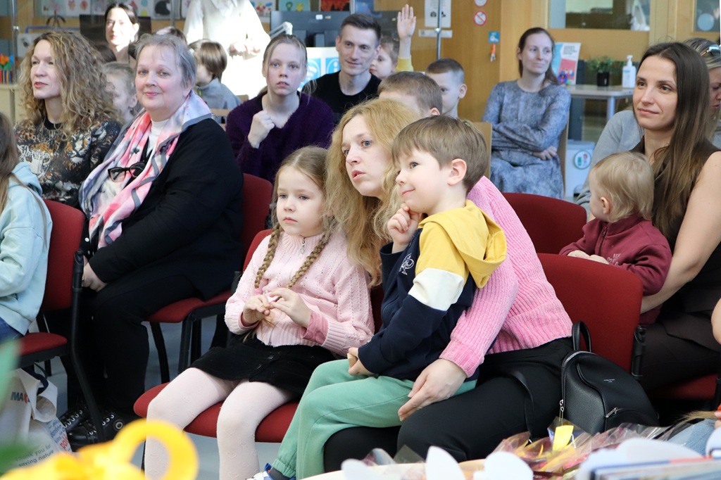 Dažāda pieaugušo un bērnu grupa uzmanīgi sēž sarkanu krēslu rindās, visi ar seju uz priekšu. Noskaņojums ir koncentrēts un iesaistīts.