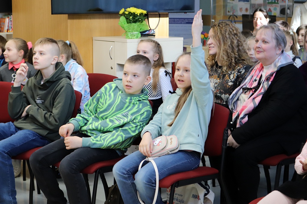 Telpa, kurā sēž uzmanīgi bērni un pieaugušie. Meitene paceļ roku, dedzīgi vēloties piedalīties. Telpu piepilda spilgtas, iesaistītas sejas izteiksmes.