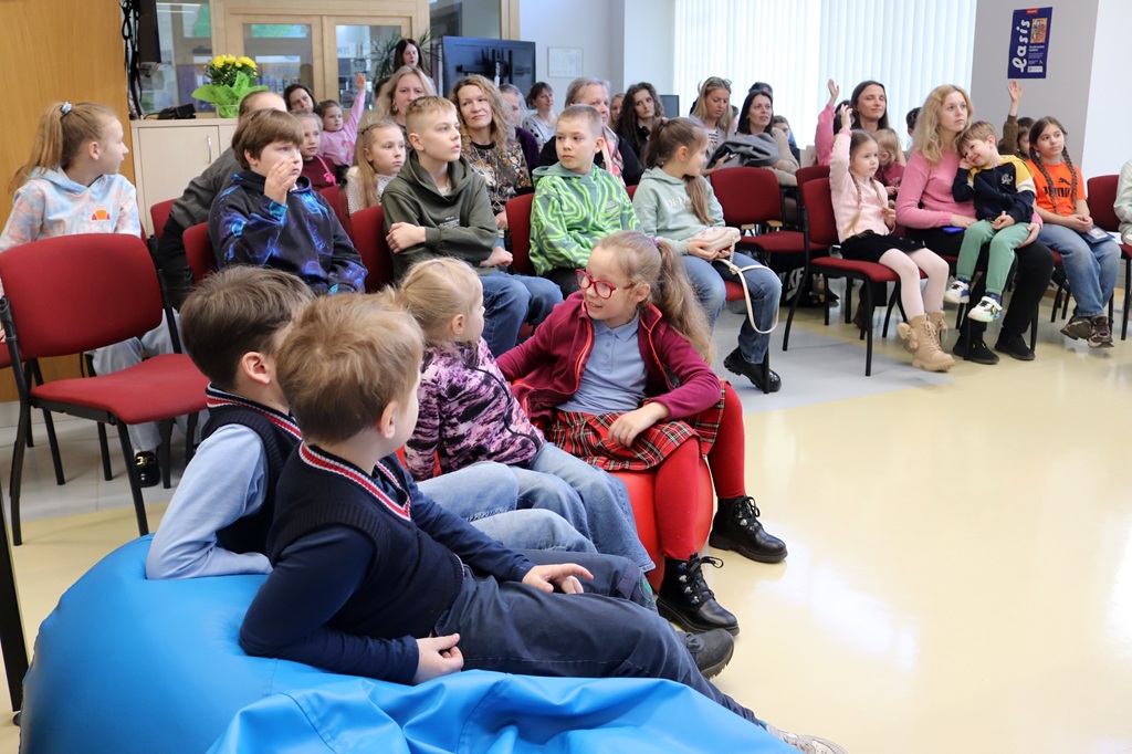 Bērnu un pieaugušo grupa sēž sarkanos krēslos spilgti apgaismotā telpā. Daži bērni ir apsēdušies uz zila pupu maisa. Atmosfēra ir uzmanīga un saistoša.