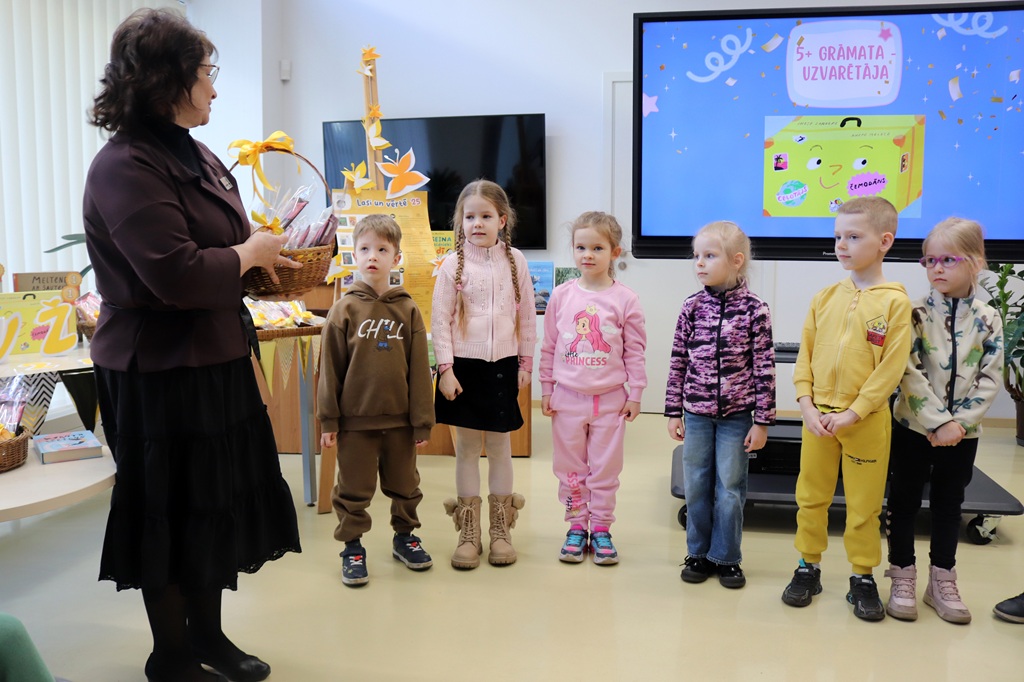 Sieviete stāv, turot rokās grozu, un uzrunā sešus mazus bērnus, kas sarindoti klasē. Krāsains ekrāns fonā attēlo rotaļīgu dizainu. Atmosfēra ir dzīvespriecīga un izglītojoša.