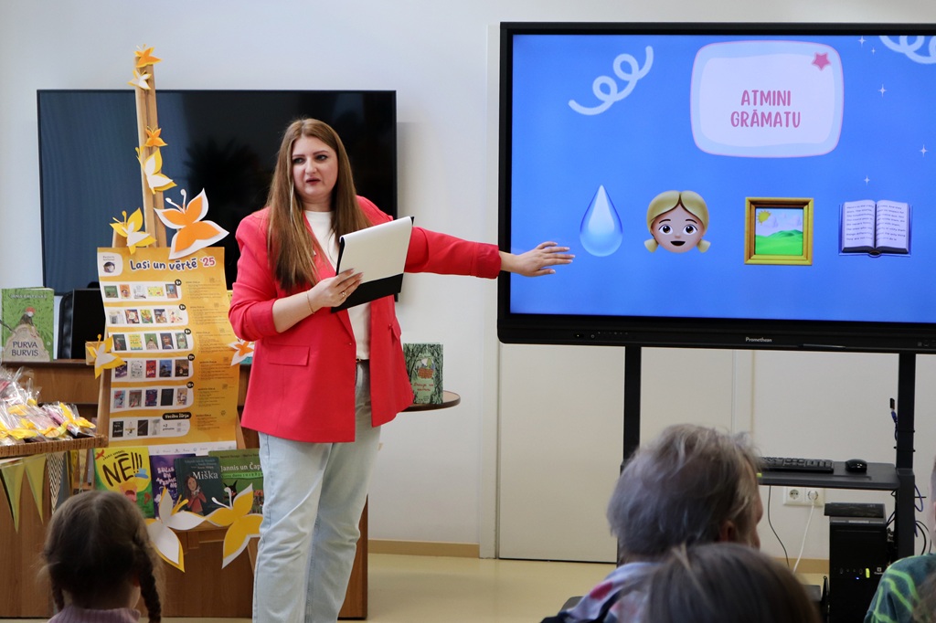 Sieviete sarkanā žaketē stāv ekrāna priekšā, kurā redzamas emocijzīmes un teksts. Auditorija uzmanīgi klausās. Telpā valda radoša, izglītojoša atmosfēra.