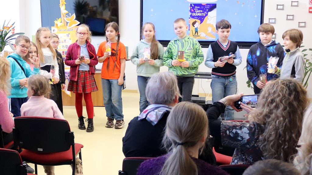 Bērnu grupa stāv rindā, turot rokās sertifikātus un dāvanas, ar skatu pret auditoriju. Viņi atrodas spilgti apgaismotā telpā ar krāsainiem rotājumiem un aiz tiem esošu ekrānu, kas liek domāt par svinīgu notikumu. Auditorija, daži fotografējas, šķiet iesaistīta un atbalstoša.