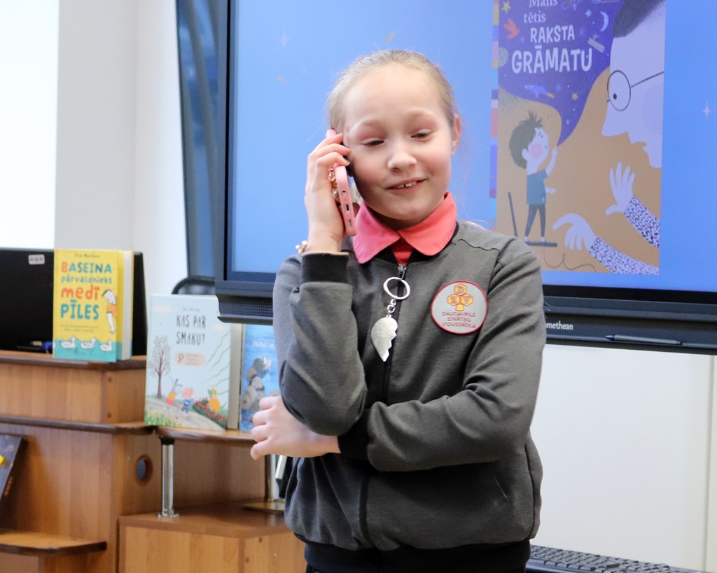 Jauna meitene pelēkā džemperī ar kakla siksniņu smaida, runājot pa tālruni. Fonā grāmatas un ekrāns attēlo krāsainas ilustrācijas.