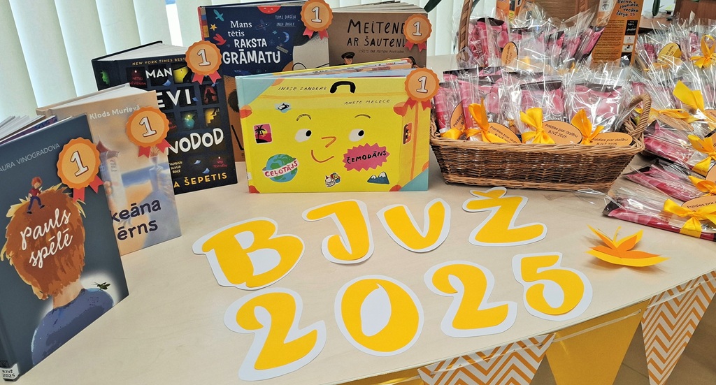 Izstādītas grāmatas ar dzeltenām etiķetēm un grozs ar našķiem. Uzraksts "BJVŽ 2025" ir izcelts ar trekniem, dzelteniem burtiem. Valda svētku atmosfēra.