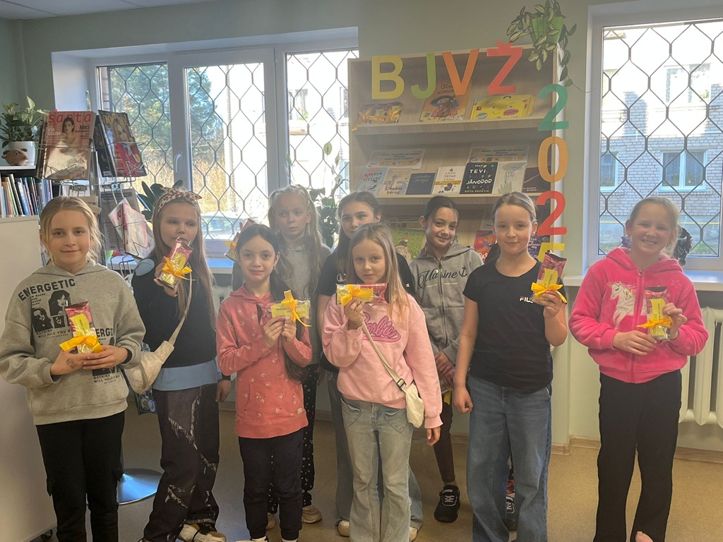Smaidīgu bērnu grupa bibliotēkā, turot dzeltenos un rozā iepakojumos iesaiņotus priekšmetus. Fonā redzami grāmatu plaukti un burti "BJVŽ 2025"