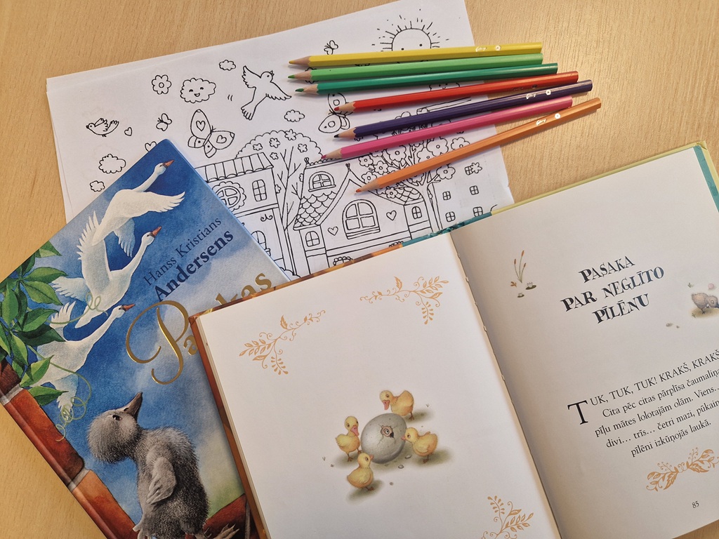 Bērnu grāmatas un krāsainie zīmuļi uz galda; ieskaitot Andersena pasakas ar gulbja vāku un atvērtu lapu ar pīlēniem, kas pauž radošumu un iztēli.