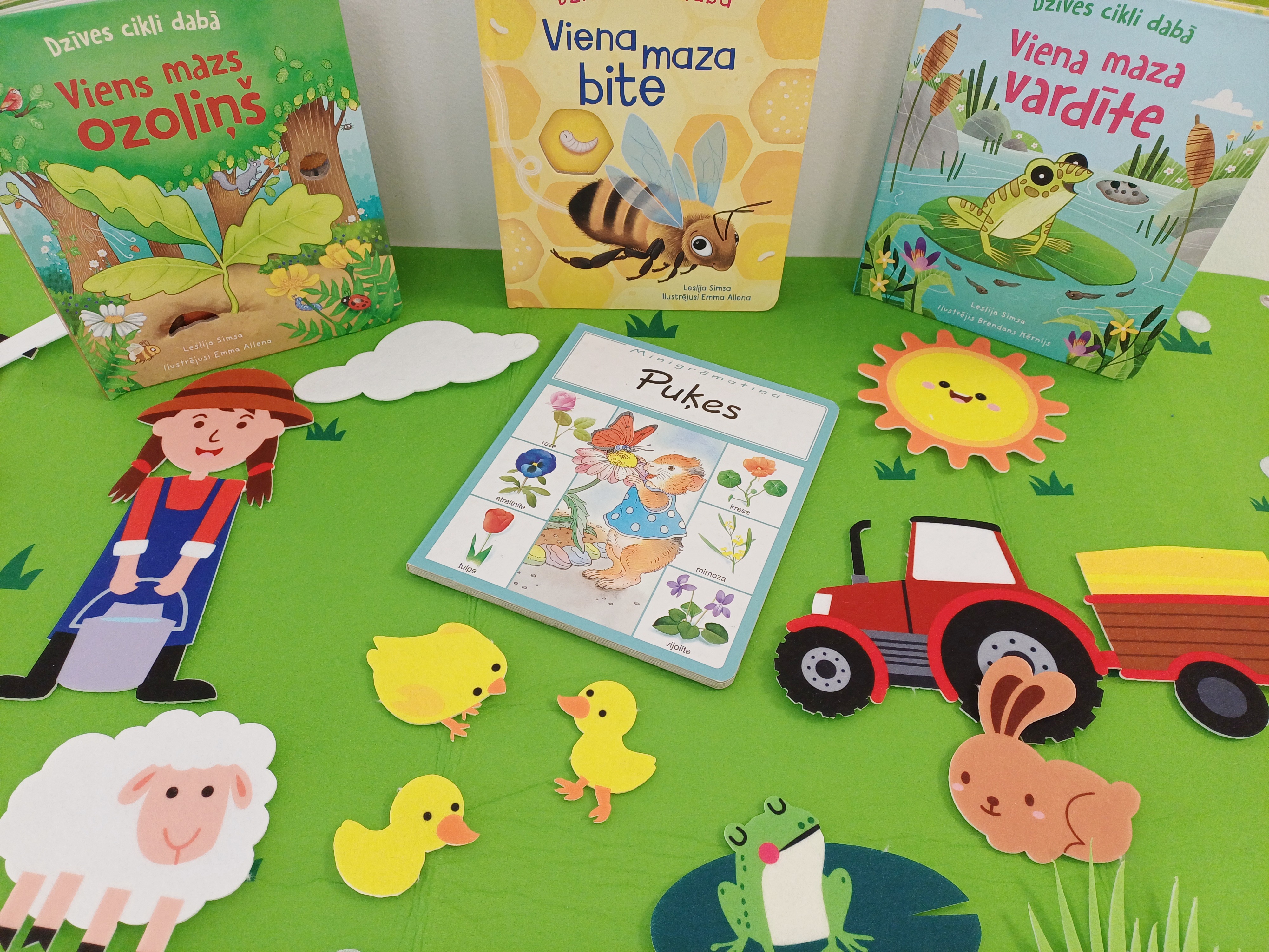 Bērnu grāmatas un izgriezumi uz zaļa fona. Grāmatās attēlots koks, bite un varde. Tās ieskauj krāsaini papīra dzīvnieki, saule un traktors. Jautrs un rotaļīgs tonis.