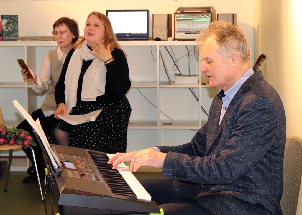 Vīrietis spēlē klavieres mājīgā istabā, kamēr divas sievietes uzmanīgi klausās netālu.