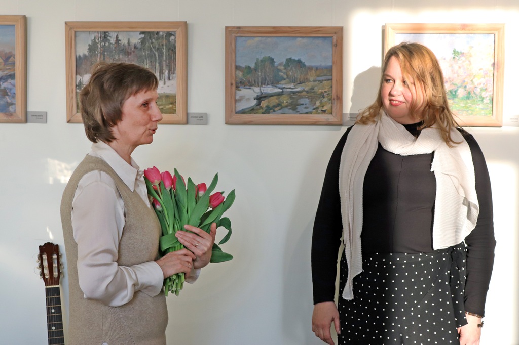 Divas sievietes stāv blakus, katra turot rokās krāsainu ziedu pušķi, un smaida kamerai.