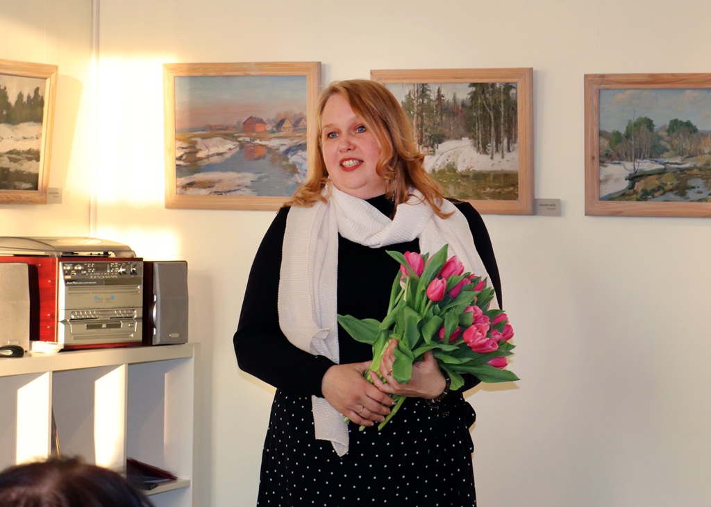 Sieviete smaida, rokās turot spilgtu ziedu pušķi.