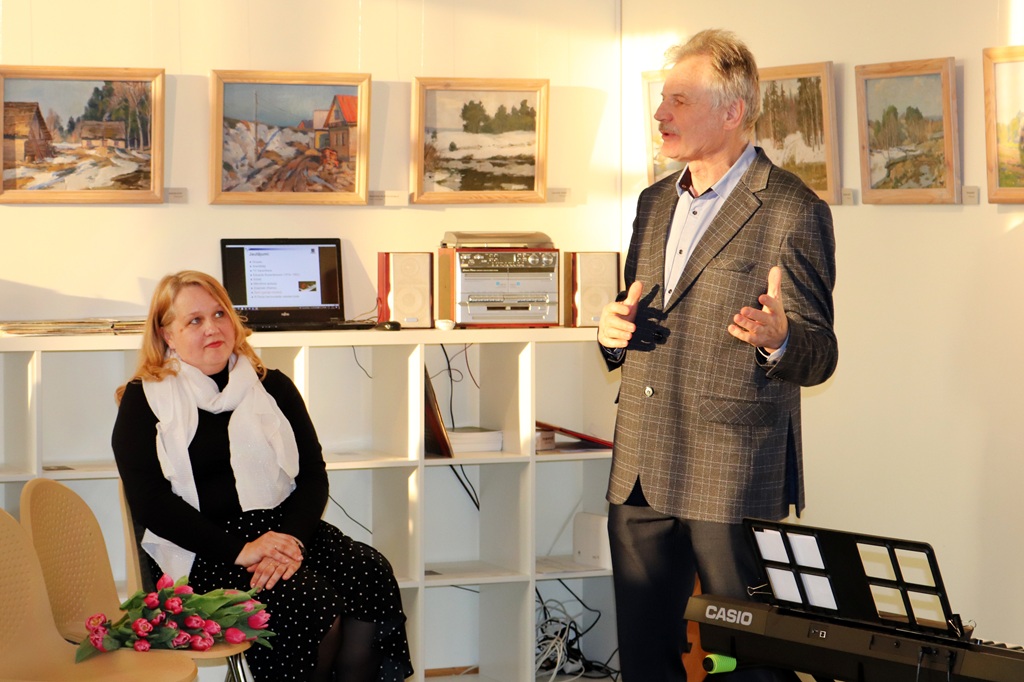 Vīrietis melnā jakā stāv pārliecināti ar neitrālu sejas izteiksmi un izplūdušu fonu.