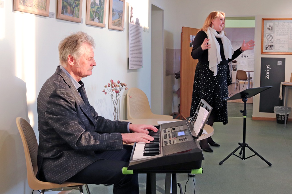 Vīrietis un sieviete priecīgi kopā spēlē mūziku uz klavierēm, viņus ieskauj silta un mājīga atmosfēra.