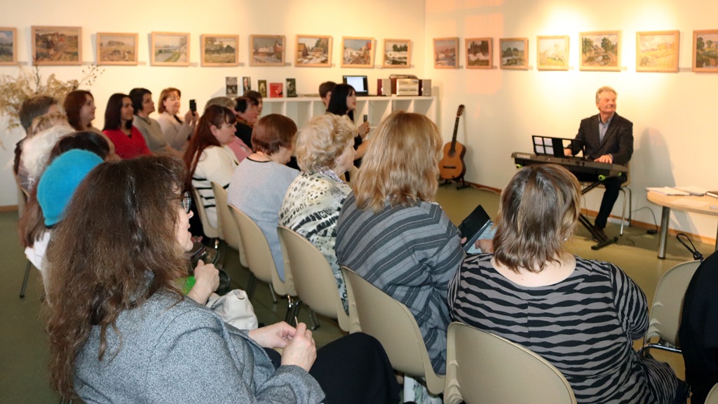Vairāki cilvēki sēž krēslos, piedaloties diskusijā plašā telpā.