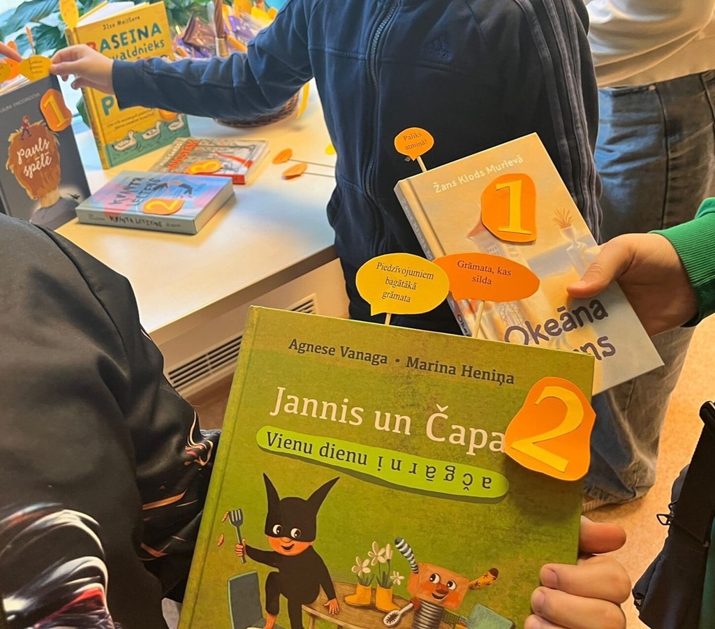 Divi cilvēki pie galda tur rokās grāmatas. Priekšplānā esošā grāmata ar nosaukumu 'Jannis un Čapa, Vienu dienu ačgarni' ir atzīmēta ar lielu, oranžu ciparu '2'.