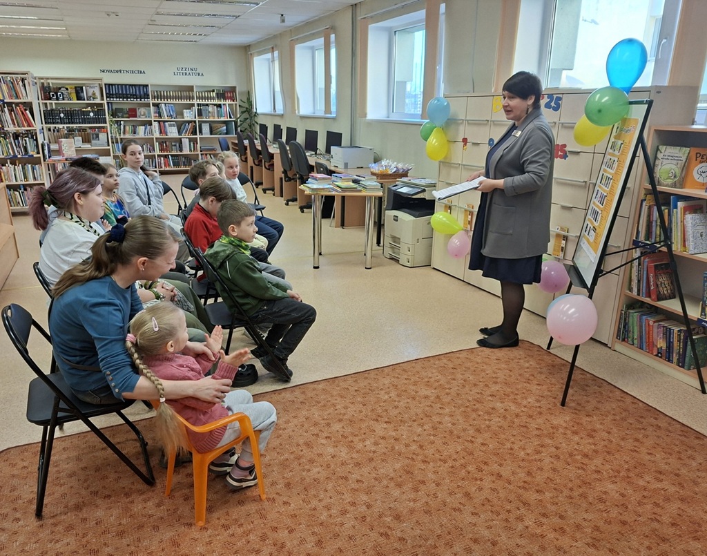 Bērnu un pieaugušo grupa bibliotēkā sēž uz krēsliem, kamēr viņu priekšā stāv bibliotekāre ar grāmatu rokās. Telpa ir dekorēta ar krāsainiem baloniem, un pie sienām izvietoti grāmatu plaukti, radot mājīgu un aicinošu atmosfēru.