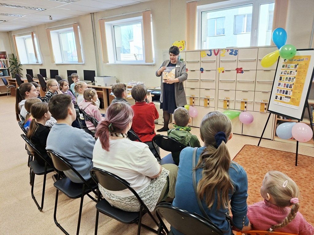 Bibliotekāre bibliotēkā rāda grāmatu sēdošu bērnu grupai. Koši baloni un rotājumi rada svētku noskaņu, raisot prieku un ieinteresētību.
