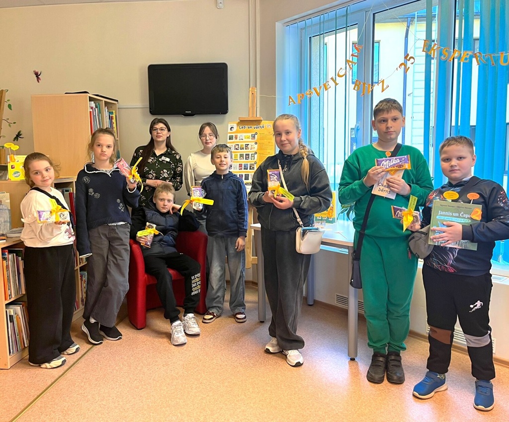 Smaidīgu bērnu un pieaugušo grupa stāv bibliotēkā, turot rokās grāmatas un mazus saiņojumus. Fonā redzami grāmatu plaukti un logs.